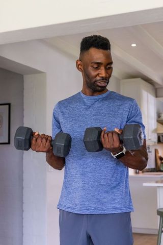 African american man lifting dumbbells in kitchen wearing smartwatch and blue shirt