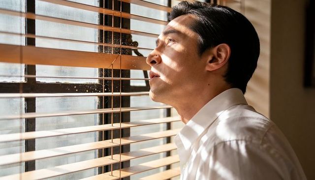 Man in white shirt peering through wooden blinds bathed in warm striped sunlight