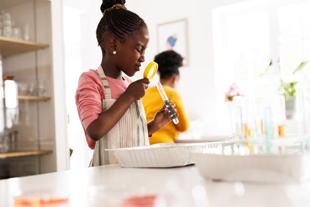 This image depicts a young African American girl in a kitchen, examining a test tube with a magnifying glass. It is perfect for illustrating concepts related to childhood curiosity, learning, and science education. Ideal for use in educational materials, websites promoting STEM activities for children, or advertisements for educational toys and kits.