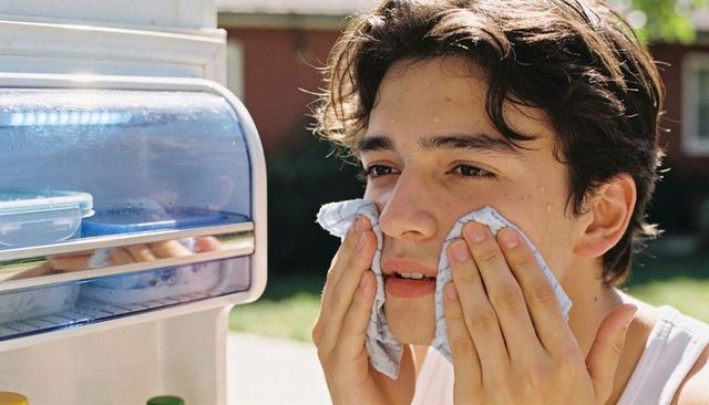 Teen male cooling face with cloth by cooler in sunlit backyard for summer relief skincare