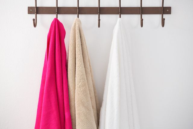 Three towels in pink, beige, and white hanging on a brown hook rack against a white wall. Ideal for use in articles or advertisements related to home decor, bathroom organization, cleanliness, and household tips.