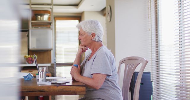 Senior woman in gray casual attire using laptop at home, sitting at a wooden table in a bright room with large windows. Ideal for illustrating themes like senior technology use, remote work, elderly independence, and active aging.