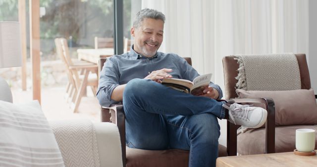 Middle-aged man sitting on a comfortable chair in a living room, casually dressed, with a book in hand and smiling. Ideal for concepts of leisure, relaxation, reading habits, home lifestyle, and alone time. Could be used for advertisements, magazines, or websites focusing on self-care, home living, and leisure activities.