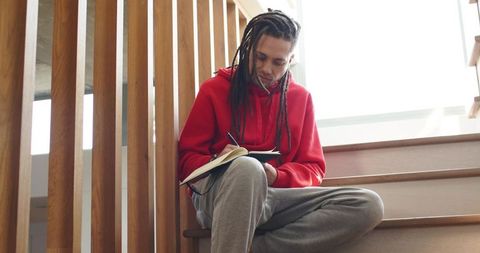 African american male writing in notebook on stairs wearing red hoodie, natural light