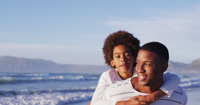 Father giving son piggyback ride on scenic beach with ocean in background. Ideal for depicting family bonding, carefree summer moments, and outdoor activities. Useful in marketing materials for family-oriented products, travel destinations, or parenting articles.