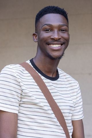 Confident young man smiling in striped tee with crossbody strap on neutral urban backdrop