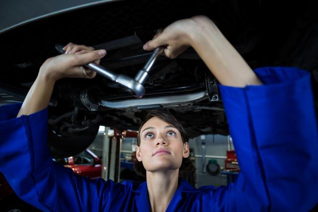 Image shows a female mechanic repairing a car in a garage while wearing a blue uniform and using a wrench. Useful for content promoting gender diversity in skilled trades, automotive repair services, mechanical training programs, and workshop advertisements.