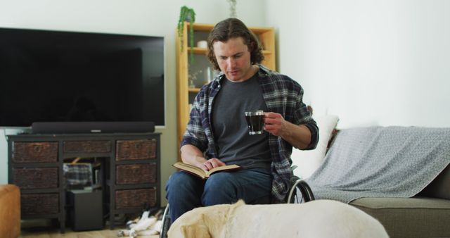 Relaxed caucasian disabled man in wheelchair reading book and drinking coffee in living room. domestic lifestyle with physical disability.