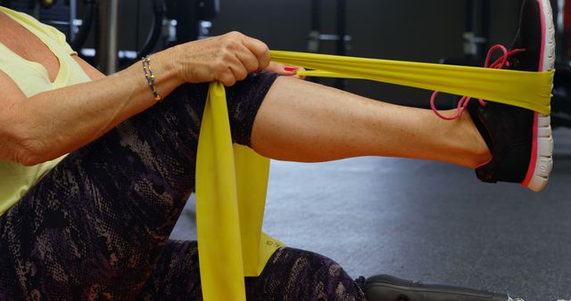 Older adult using a yellow resistance band for rehabilitation exercises in a gym. Perfect for illustrating physical therapy sessions, senior fitness, and health-related content. Useful for brochures or websites focused on senior wellness, rehabilitation programs, or resistance band workouts.