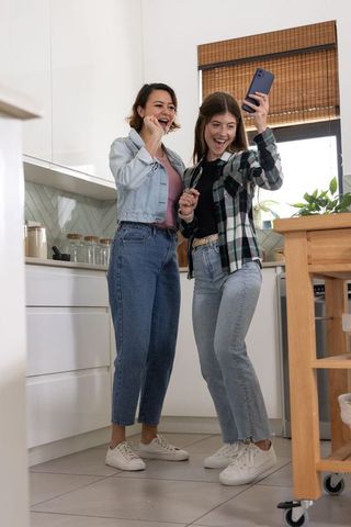 Friends posing for selfie in bright modern kitchen, holding smartphone and laughing together