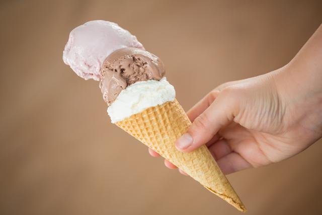 This close-up of a hand holding a waffle cone with three scoops of different ice cream flavors is perfect for illustrating concepts of indulgence, summer treats, and dessert enjoyment. It is ideal for use in food blogs, restaurant menus, advertisements for ice cream parlors, and summer-themed marketing campaigns.