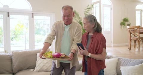 Senior couple sharing fruit platter in sunlit living room offering fresh tropical snack and companio