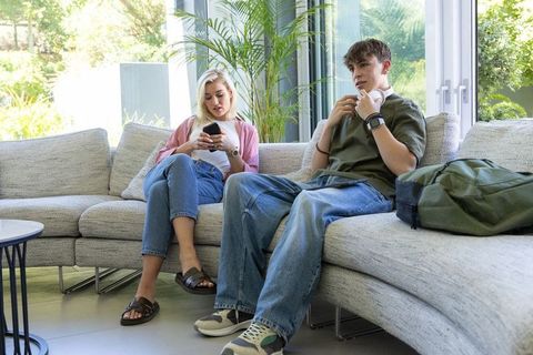Teenager and woman relaxing on sofa while scrolling smartphone and listening to music