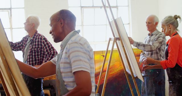 Senior adults are actively participating in a group art class, painting on canvases with easels. Bright, naturally lit studio enhances the creative and positive atmosphere. Ideal for use in articles about senior activities, lifelong learning, creativity during retirement, and community engagement.