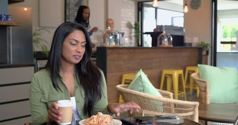 Indian woman sitting in cozy coffeehouse sipping takeout coffee and reaching for phone