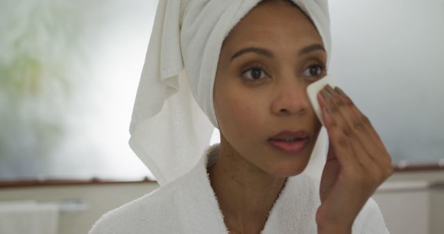 Woman wearing white bathrobe and towel on head applying skincare product on face in bright bathroom. Useful for articles or ads related to self-care routines, skincare products, beauty tips, health and wellbeing.