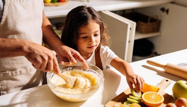 Parent stirring batter while child grabbing sliced kiwi and orange on kitchen counter