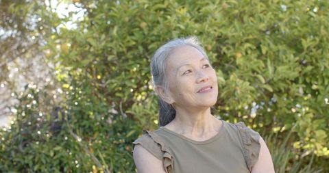 Senior asian woman standing gazing upward in sunlit garden, serene mature portrait