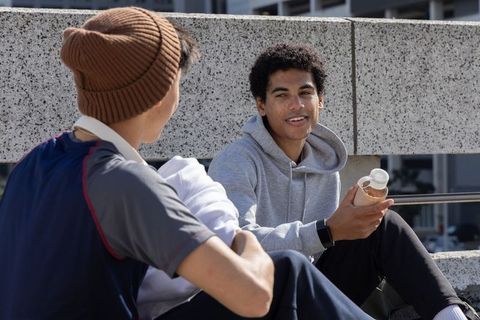 Multiracial teen friends chatting on urban ledge holding water bottle and wearing hoodies