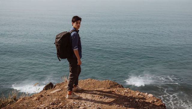 Backpacker standing on coastal cliff gazing toward camera with ocean view and backpacking gear