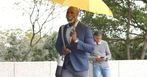Senior african american man walking with yellow umbrella and smartphone in urban park