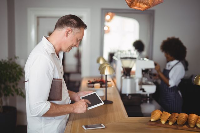Man using digital tablet at restaurant counter, holding notebook. Barista working in background. Ideal for themes related to modern business, technology in hospitality, customer service, and professional environments.