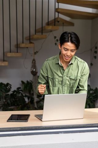 Young asian man cheering at laptop in modern loft with greenery and wooden staircase