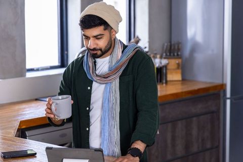 Young man working remotely in modern apartment kitchen holding coffee mug and tablet