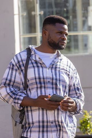 African commuter checking smartphone on sunny urban sidewalk in plaid shirt and backpack