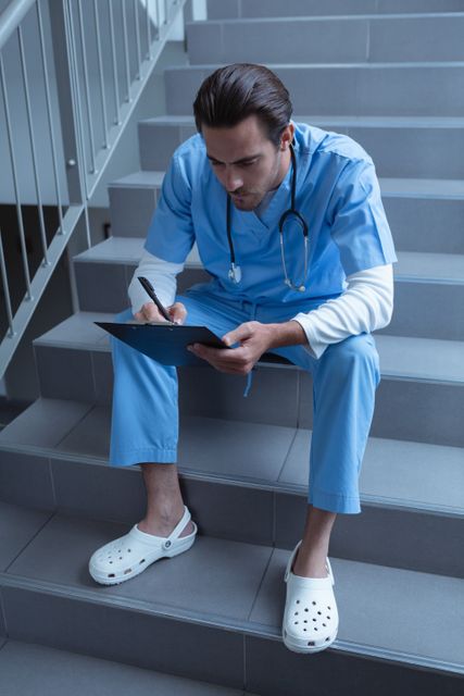 High angle view of male surgeon writing on clipboard while sitting on stairs at hospital