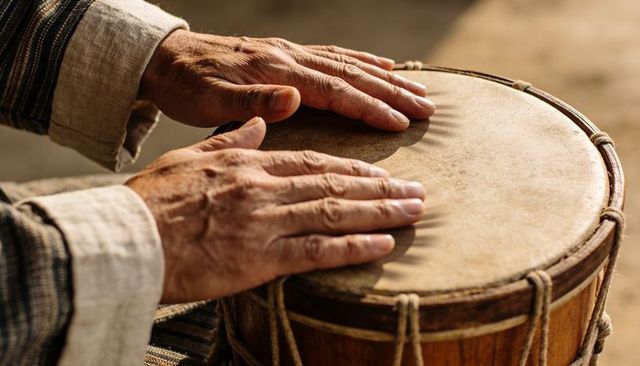 Sunlit senior hands playing rope-tuned wooden hand drum, close-up rustic percussion