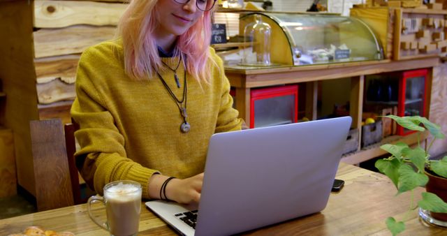 Young woman with pink hair typing on laptop in cozy coffee shop. Coffee and plants on wooden table. Suitable for concepts like freelancing, remote work, young professional, casual work environment, cozy cafe atmosphere.