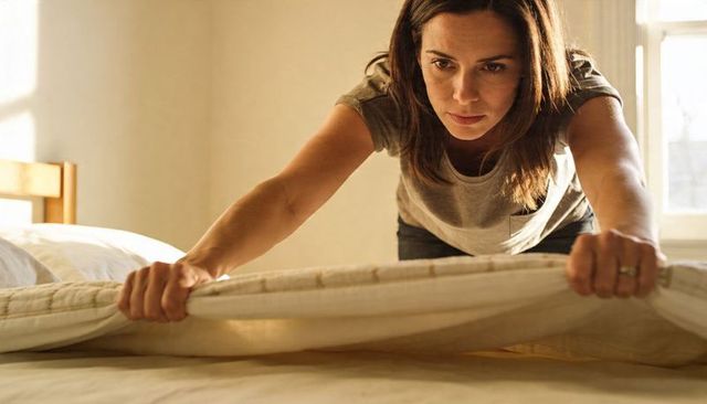 Woman making bed in sunlit minimalist bedroom leaning forward, tucking fitted sheet