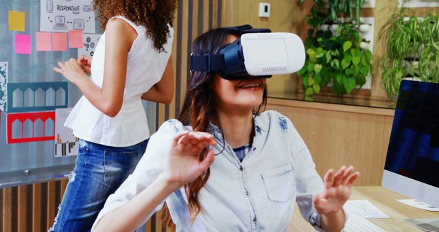 Woman engaged with virtual reality in a modern, casual office setting. Vibrant workspace with plant decor and colleague working at a whiteboard. Ideal for showcasing innovative work environments, technology integration, and interactive professional settings.