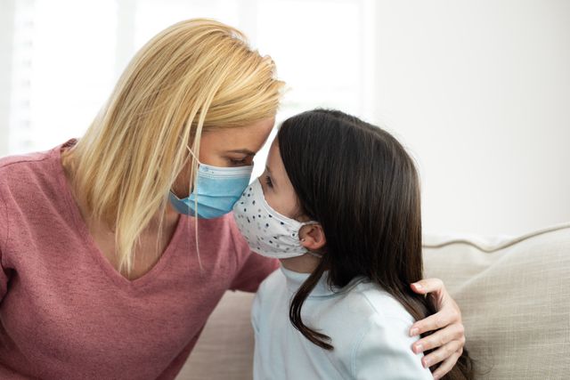 Caucasian mother and daughter sitting on couch at home, pressing faces together while wearing facemasks. Ideal for use in articles or advertisements about family bonding during the pandemic, health and safety measures, or emotional connections in challenging times.