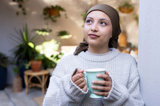 Happy biracial woman in hijab holding coffee, looking away in living room at home, copy space. Happiness, health, inclusivity and domestic life.