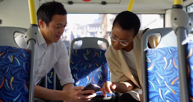 Two professionals are engaging in a discussion on a city bus, showing a moment of urban commuting while collaborating. They are looking at a mobile device, possibly reviewing work materials. This scene can be used to represent business interactions, daily commuting, teamwork, or any context of professional discussion on the go.