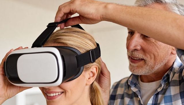 Senior man adjusting vr headset on smiling woman at home trying virtual reality