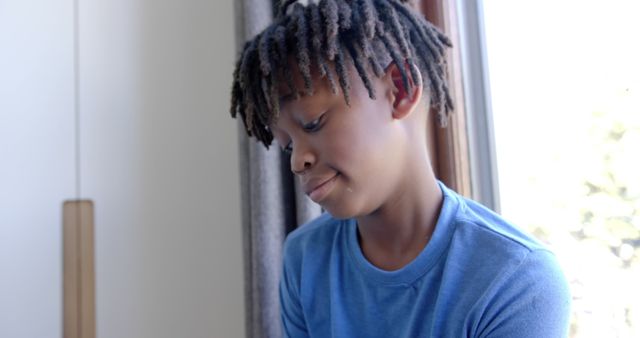 Depicting a young Black boy with dreadlocks, wearing a blue shirt, looking thoughtful indoors by a window with natural light. Ideal for diverse representation in educational content, emotional and introspective themed materials, or advertisements focusing on peaceful moments and childhood.
