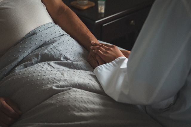 Senior man lying in bed holding hands with a medical worker, symbolizing support and compassion. Ideal for use in healthcare, home care, and elderly care contexts to depict patient support, medical assistance, and compassionate care.