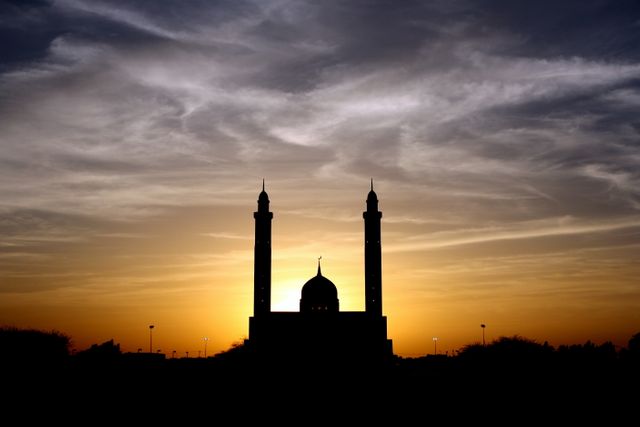 Silhouette of a mosque with twin minarets against a vibrant sunset sky. Ideal for themes of spirituality, Middle Eastern culture, Islam, architecture, peace, and tranquility. Useful in articles, presentations, and posters related to religious festivals, cultural diversity, and evening landscapes.