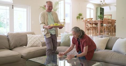 Senior couple carrying tray and placing drinks on glass coffee table in sunlit living room