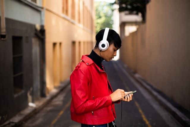 Side view of a fashionable biracial transgender in the street, walking on the streets, listening to music on headphones, using smartphone
