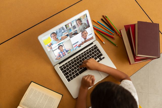 Child attending an online class or meeting with other students through a laptop. Colorful pencils and books around signify a study environment. Useful for illustrating online education, virtual classrooms, remote learning setups, and digital communication among students.