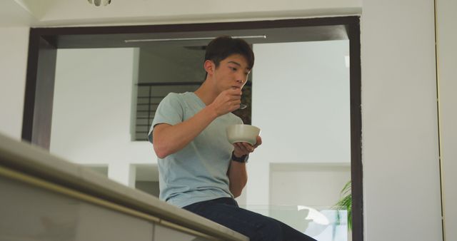 Young man in casual attire enjoying breakfast in a modern kitchen. Great for use in articles or advertisements related to healthy living, morning routines, kitchen designs, or home life.