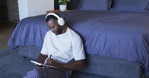 Middle aged black man sitting on bedroom floor writing in notebook while wearing headphones