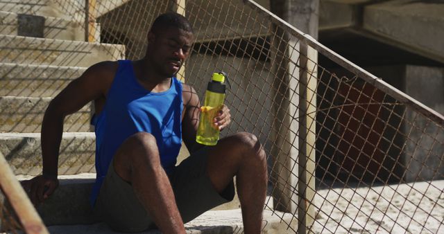 An exhausted male athlete in sportswear is sitting on a staircase, holding a water bottle, and catching his breath. The stairway appears to be outside, potentially in an urban setting. This can be used in contexts about fitness, workout recovery, the importance of hydration, or perseverance in sports.