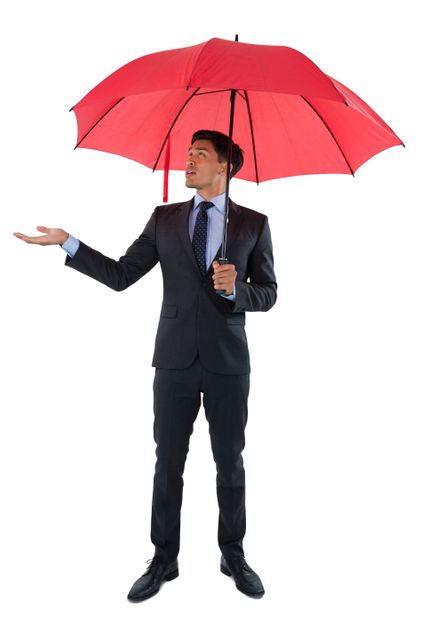 Businessman in a suit holding a red umbrella, standing against a white background. Ideal for concepts related to protection, corporate environment, weather preparedness, and professional attire. Suitable for use in business presentations, advertisements, and articles on professional conduct.