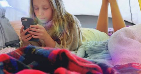 Lying girl holding smartphone inside cozy blanket fort, scrolling and relaxing at home