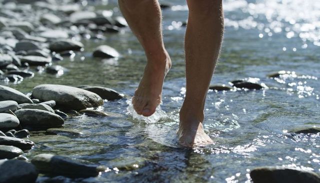 Wading barefoot through sunlit rocky stream with clear water ripples and pebble reflections